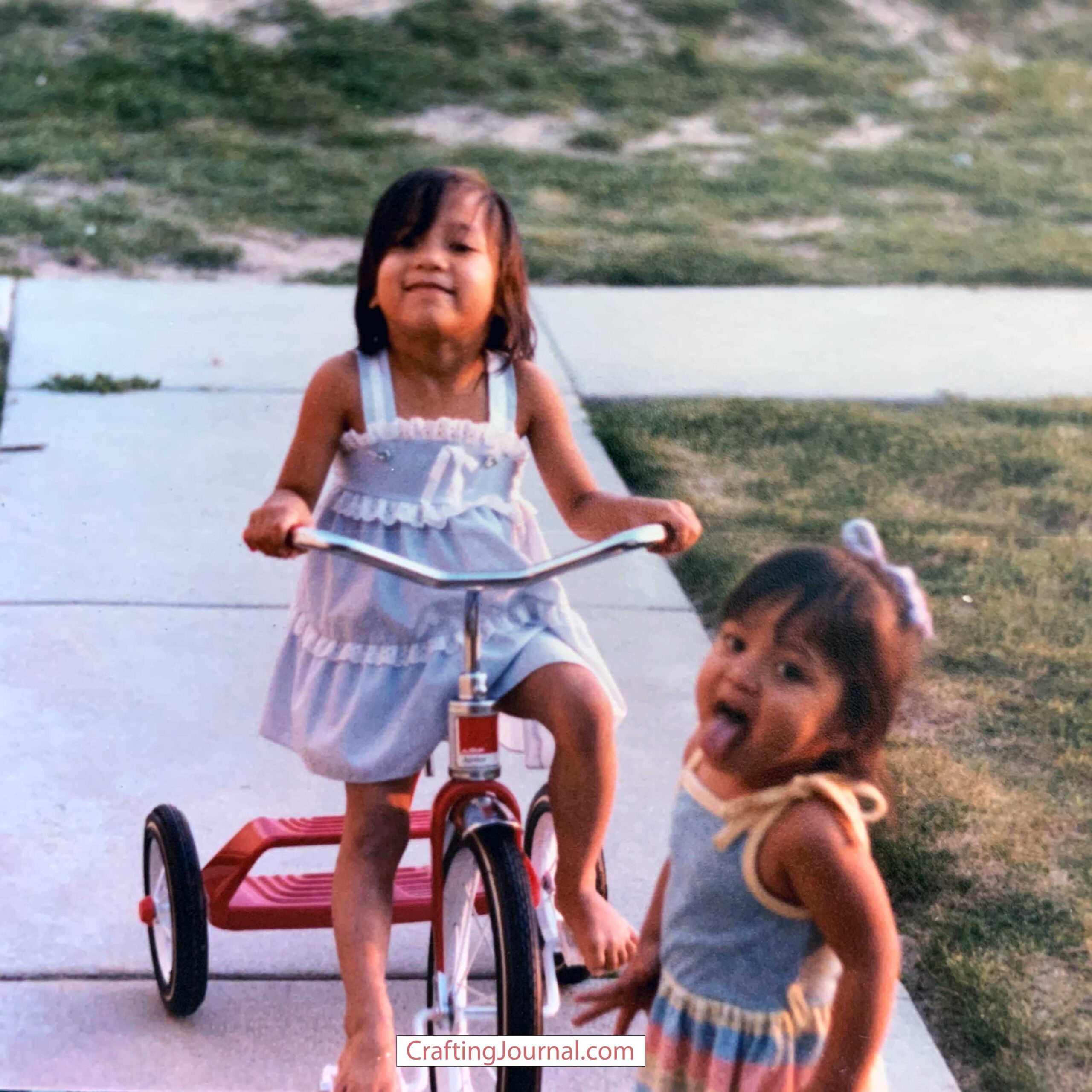 Two children playing outside on a sidewalk. One is on a red tricycle and the other is standing nearby